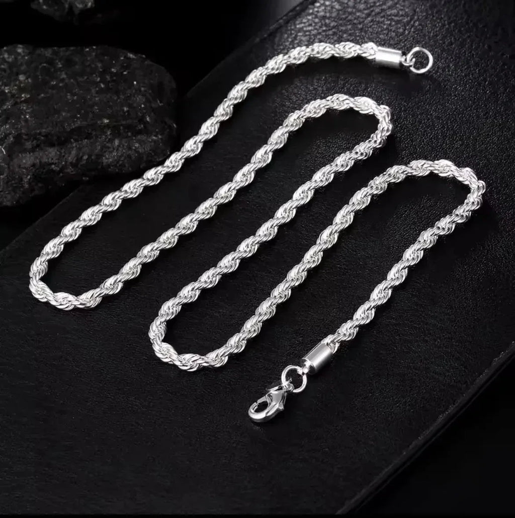 "Die Halskette „Helga“ aus Silber und das passende Armband mit Karabinerverschluss von flyliving präsentieren sich auf einer strukturierten schwarzen Oberfläche, akzentuiert durch schwarze Steine im Hintergrund."