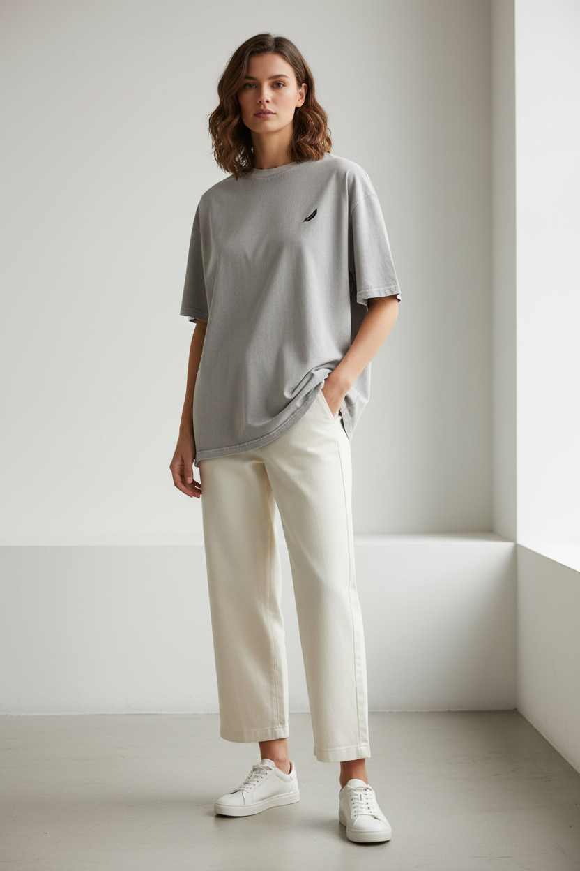 Oversized unisex grey T-shirt by flyliving with a small black feather-like logo on the chest, worn by a woman with hands in pockets, styled with light beige wide-leg pants and white sneakers.