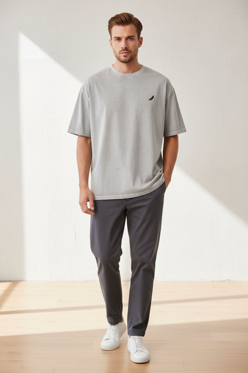 Washed grey oversized t-shirt by flyliving with a small black feather logo on the chest, worn by a male model with dark trousers and white sneakers.