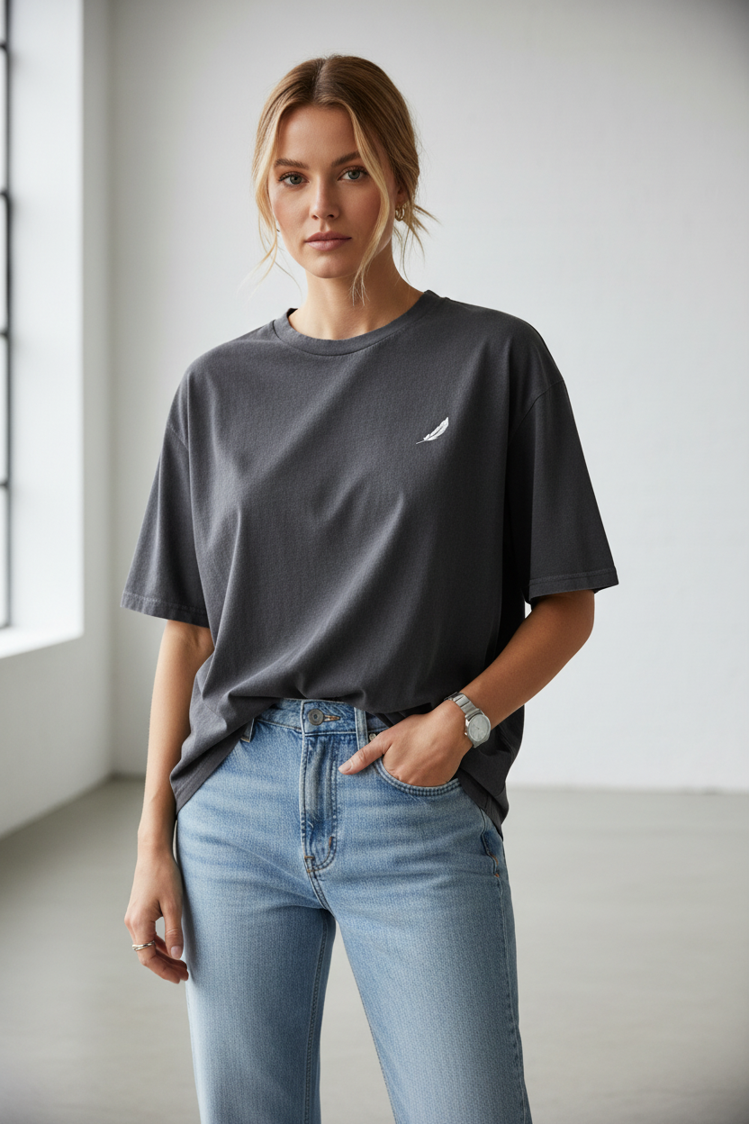 Dark grey oversized t-shirt with a small white feather logo by flyliving, worn by a woman in light wash jeans and a silver watch.