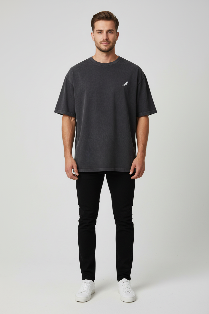Oversized dark grey T-shirt by flyliving with a small white curved logo on the chest, worn by a male model with black jeans and white sneakers.