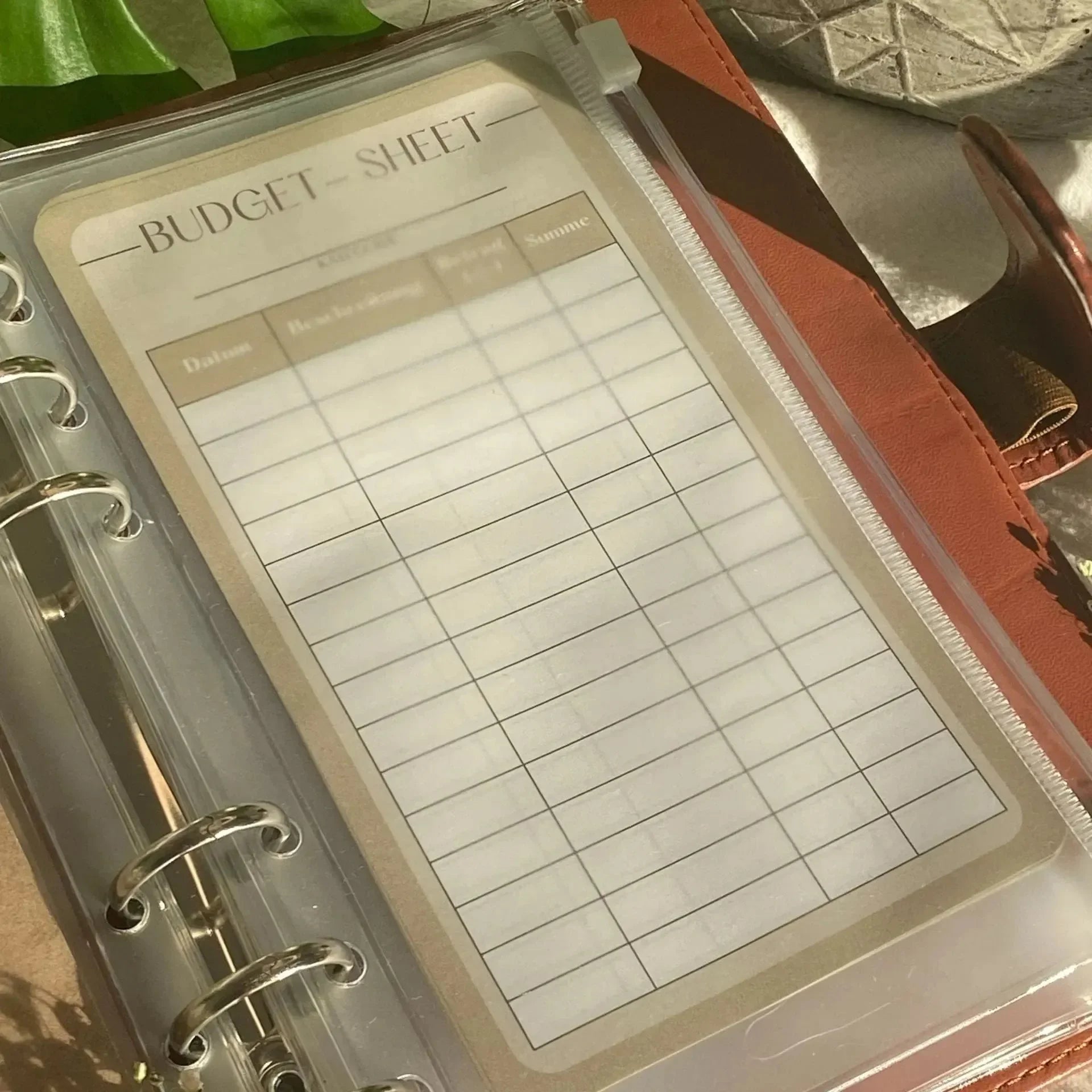 Eine Nahaufnahme von BeginnerBudget Budget Sheets (10er-Pack) in einem Ringbuchplaner mit Spalten für Datum, Beschreibung, Kategorie und Betrag, plus einem Stift - ideal für den Start Ihres Sparplans oder Haushaltsbuchs. Sonnenlicht und Pflanzenblätter vervollständigen die Szene.