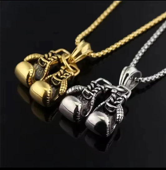 Zwei flyliving Halsketten „Angela“ mit Boxhandschuh-Anhänger, unisex und aus robustem Edelstahl, in Gold und Silber, liegen auf einer glänzenden schwarzen Oberfläche.