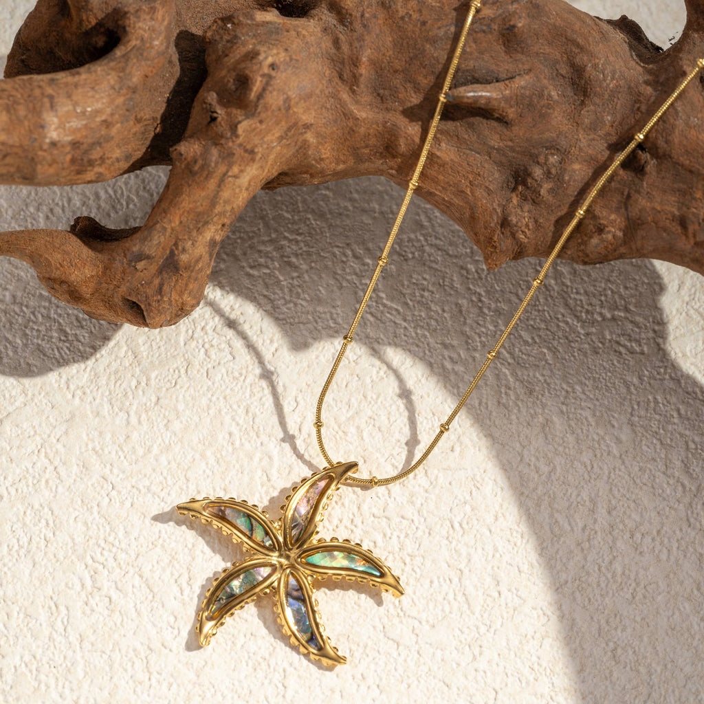 Gold-toned stainless steel starfish necklace with iridescent mother-of-pearl inlay and beaded chain by flyliving, on a textured surface with driftwood.
