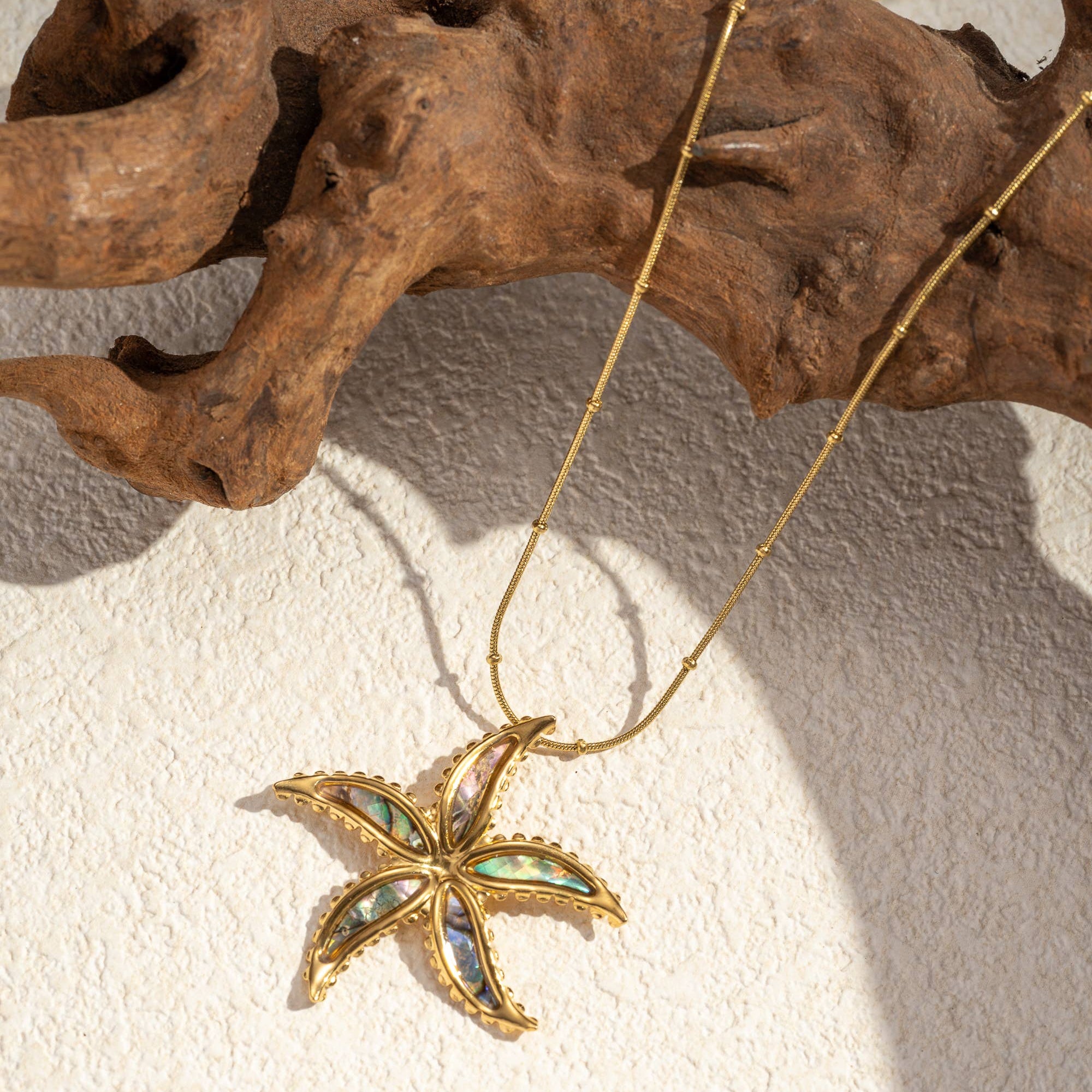 Gold-toned stainless steel starfish necklace with iridescent mother-of-pearl inlay and beaded chain by flyliving, on a textured surface with driftwood.