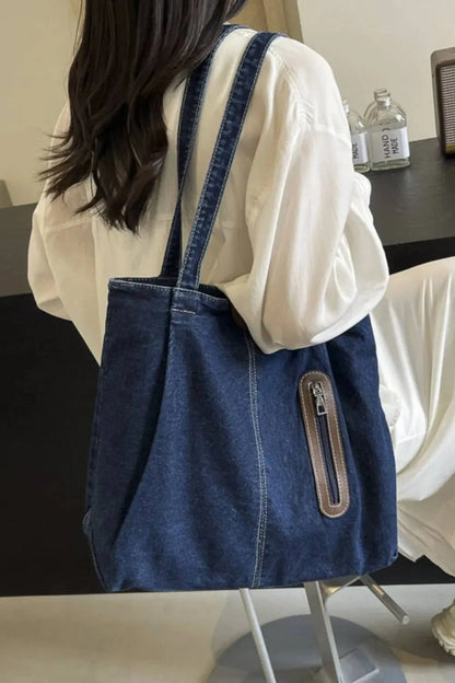 Eine Person mit langen dunklen Haaren in einem weißen Outfit sitzt auf einem Stuhl und trägt die flyliving Handtasche „Blue Jeans“, medium, für Frauen. Flaschen und ein Lautsprecher stehen auf dem Tisch dahinter.