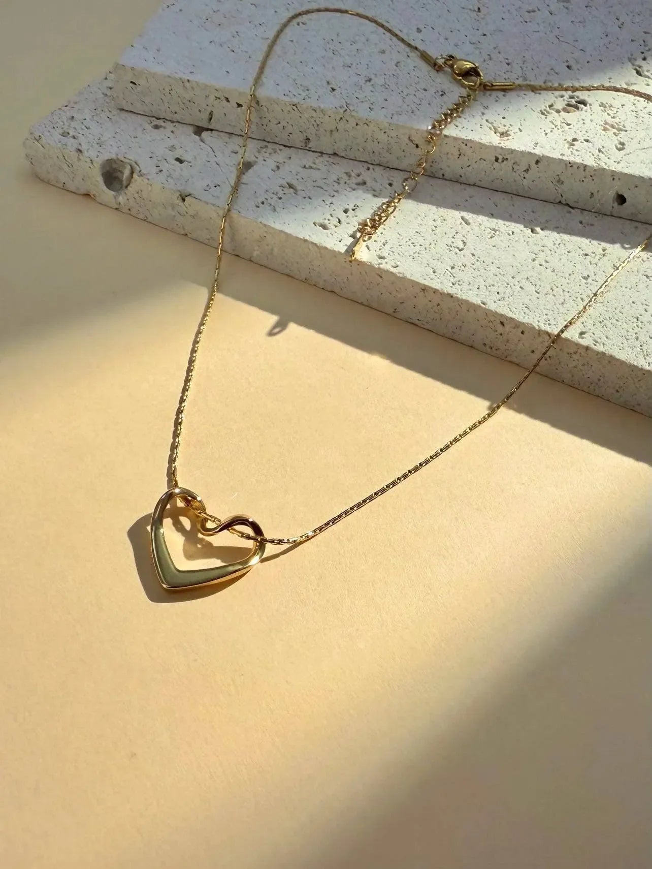 Die vergoldete Edelstahlkette mit Herzanhänger von Cosmos liegt auf einer beigen Fläche neben weißem Stein und wird sanft vom Sonnenlicht beleuchtet.