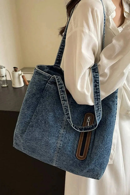 Eine Frau im weißen Hemd trägt die flyliving Handtasche „Blue Jeans“ (Medium) für Damen, die ein schickes Design mit braunen und silbernen Reißverschlussdetails aufweist. Drinnen steht sie an einem dunklen Tisch mit Teekanne und Tasse und präsentiert das dezente Markenlogo.