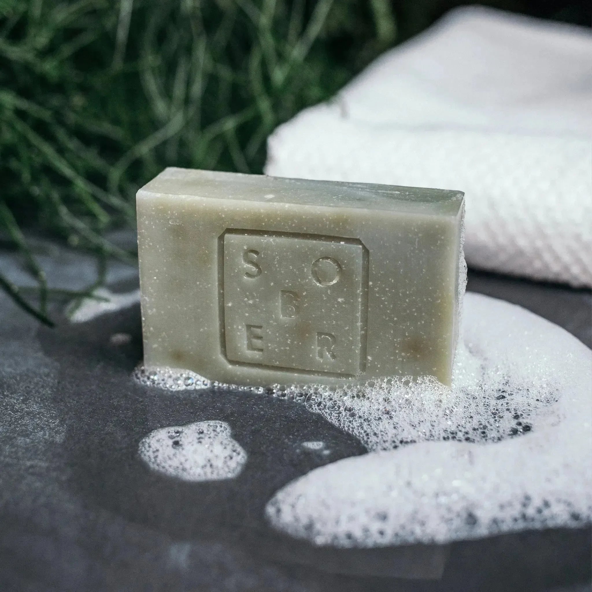 Eine Hand & Body Moisturizer Soap Bar von sober mit der Prägung "SOAP" liegt inmitten von Seifenschaum auf einer grauen Oberfläche neben einem strukturierten weißen Handtuch und grünen Blättern. Hergestellt mit veganen Ölen für eine sanfte Reinigung.