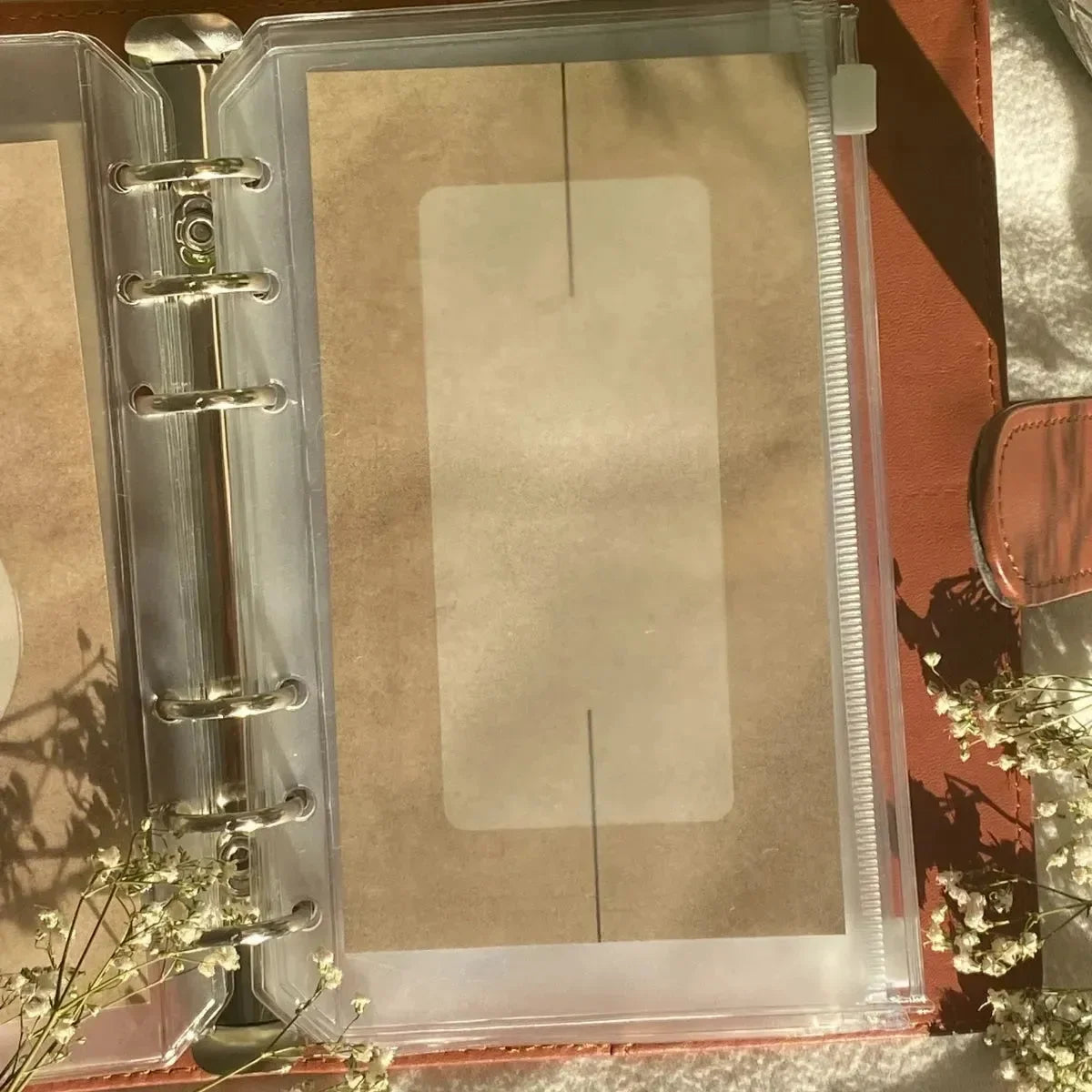 Eine Nahaufnahme eines geöffneten A6-Ordners mit braunem Einband zeigt eine BeginnerBudget Blanko Dashboards (4er-Pack) Karte in einer Plastikhülle. Sonnenlicht und weiße Blumen werfen weiche Schatten, ideal für die individuelle Beschriftung von Dashboards.