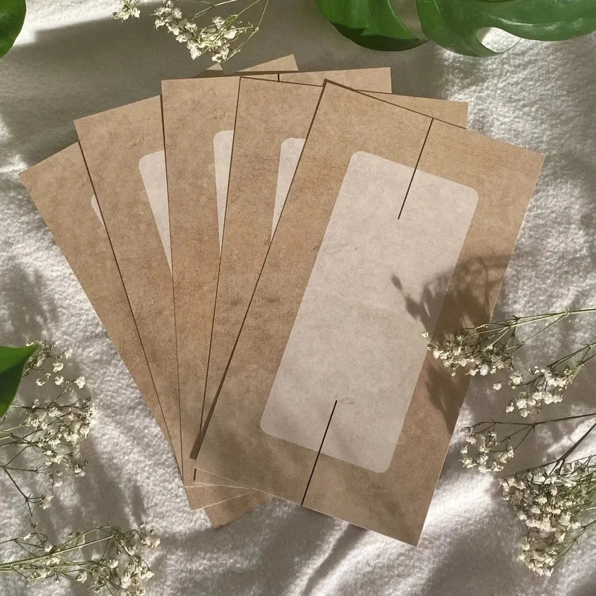 Fünf Umschläge aus dem Set BeginnerBudget Grundausstattung Budget Planung von Budget Binder sind auf seidenem Stoff aufgefächert, akzentuiert durch weiße Blumen und grüne Blätter. Jeder braune Umschlag hat ein rechteckiges Fenster für die Umschlagmethode.