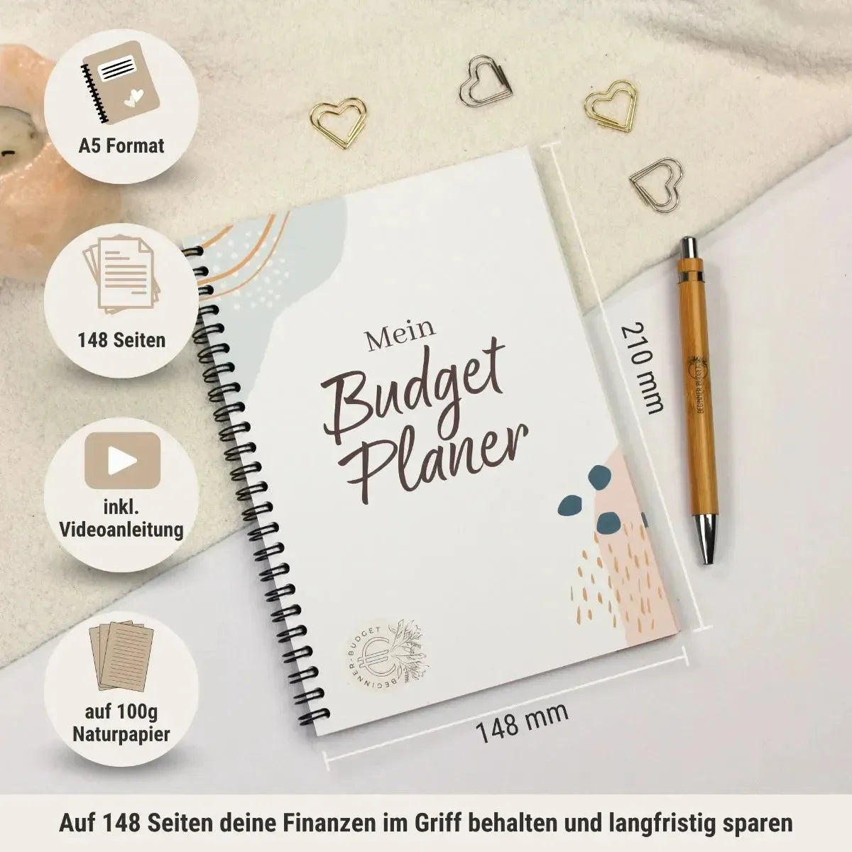 Der BeginnerBudget Budgetplaner (Jahresunabhängig) - DIN A5 mit 148 Seiten, Videoanleitung, Sinking-Funds-Teil und 100g Naturpapier ist spiralgebunden und liegt auf einem Schreibtisch neben einem goldenen Stift, goldenen herzförmigen Büroklammern und einem weißen Tuch.