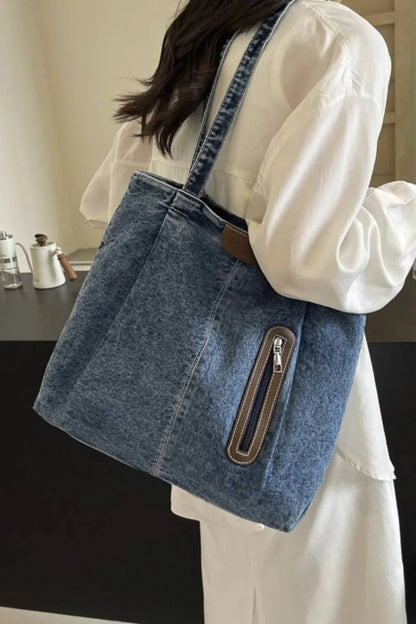 Eine Person in einem weißen Langarmkleid trägt die mittelgroße Damenhandtasche „Blue Jeans“ von flyliving mit einer Reißverschlusstasche vorne und einem braunen Etikett mit dem Markenlogo, während sie in einem Innenbereich vor einem schwarz-beigen Hintergrund steht.