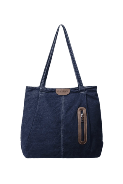 Die flyliving Handtasche „Blue Jeans“ (mittel) für Damen ist eine dunkelblaue Denim-Tragetasche mit doppelten Schultergurten, einem braunen Logo-Patch in Lederoptik und einer vertikalen Reißverschlusstasche vorne mit braunem Rand.