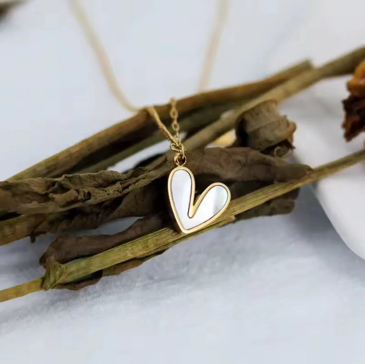 Die goldene Halskette „Uschi“ von flyliving besteht aus einem weißen, herzförmigen Anhänger mit Muschelfüllung, der auf getrockneten braunen Stielen vor hellem Hintergrund präsentiert wird.