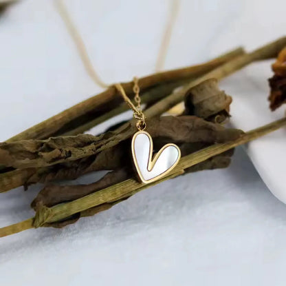 Die goldene Halskette „Uschi“ von flyliving besteht aus einem weißen, herzförmigen Anhänger mit Muschelfüllung, der auf getrockneten braunen Stielen vor hellem Hintergrund präsentiert wird.