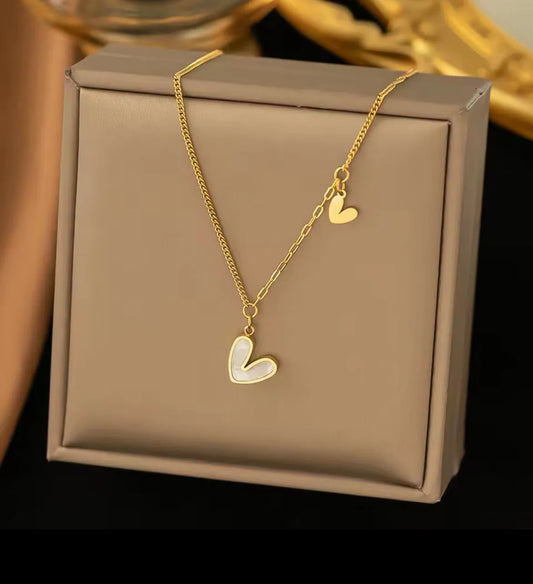 Die goldene Halskette „Uschi“ von flyliving besteht aus zwei herzförmigen Anhängern aus Edelstahl, die elegant auf einer beigen Schmuckschatulle präsentiert werden – das größere Herz in der Mitte, das kleinere hängt zart an der Seite der Kette.