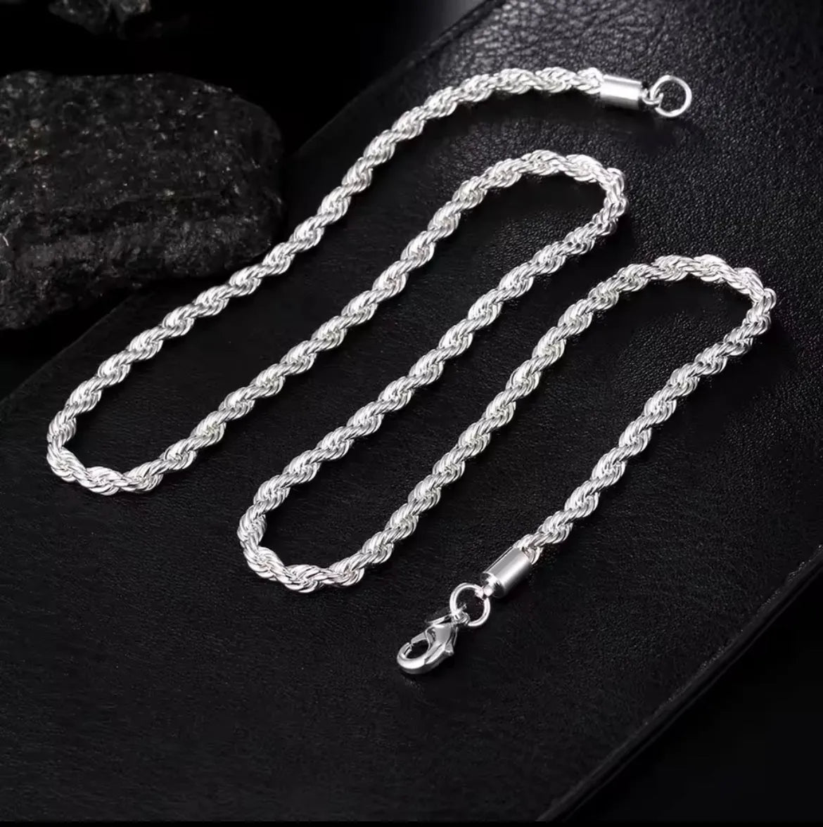 Die Halskette „Helga“ aus Silber und das passende Armband mit Karabinerverschluss von flyliving präsentieren sich auf einer strukturierten schwarzen Oberfläche, akzentuiert durch schwarze Steine im Hintergrund.