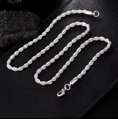 Die Halskette „Helga“ aus Silber und das passende Armband mit Karabinerverschluss von flyliving präsentieren sich auf einer strukturierten schwarzen Oberfläche, akzentuiert durch schwarze Steine im Hintergrund.