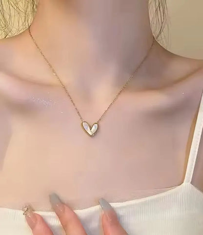 Eine Person trägt die goldene Halskette „Uschi“ von flyliving – eine herzförmige Halskette mit Anhänger – mit einem weißen Camisole-Oberteil, langen manikürten Nägeln und dezentem Schimmer auf der oberen Brust.