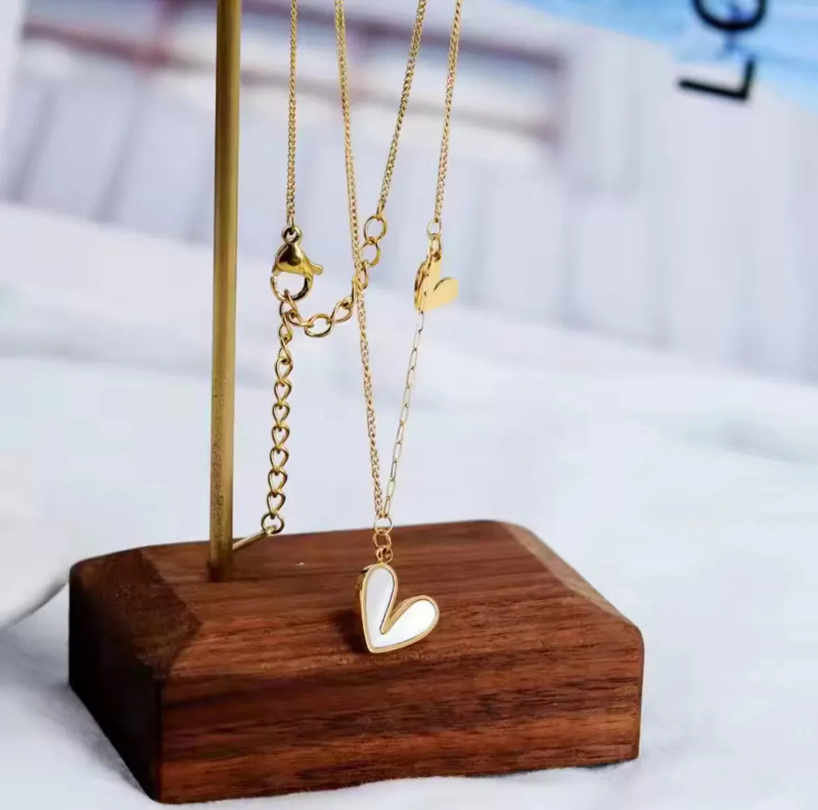 Die goldene Halskette „Uschi“ von flyliving besteht aus einem weißen herzförmigen Anhänger mit Muschelfüllung und einem kleinen goldenen Charm, präsentiert auf einem Metallständer mit Holzsockel vor einem sanft verschwommenen Hintergrund.