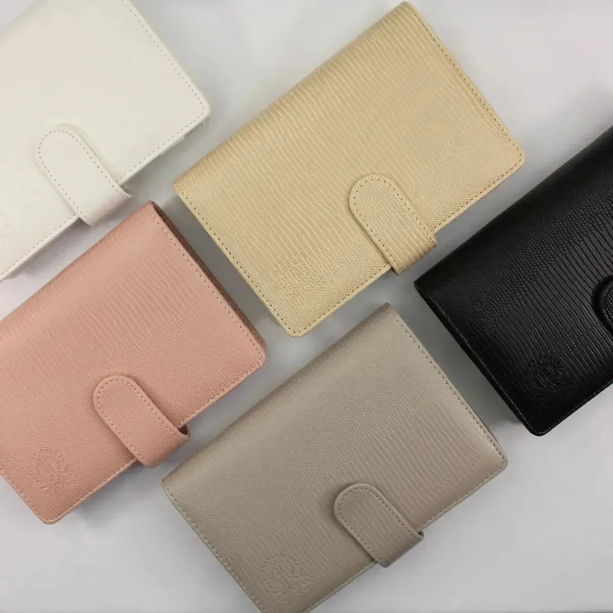 Fünf Budget Binder "Gecko" von BeginnerBudget in den Farben Weiß, Beige, Pink, Taupe und Schwarz sind auf einer hellen Fläche angeordnet - jeweils mit Druckknopfverschluss und dezenter Logoprägung - ideal für die Organisation Ihrer Finanzübersicht oder Sparziele.