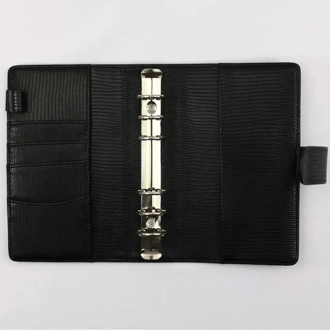 Der BeginnerBudget Budget Binder "Gecko" - Personal (A6) ist ein schwarzer, strukturierter Lederbinder mit einem Sechsring-Mechanismus, drei linken Innentaschen und einer Stiftschlaufe, offen auf einer weißen Oberfläche dargestellt.