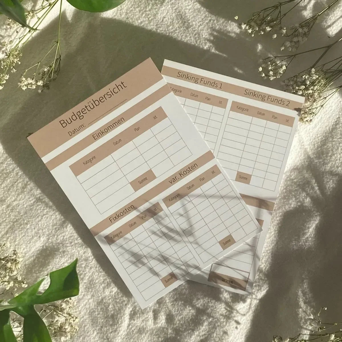 Drei deutsche Budgetblätter aus der "Grundausstattung Budget Planung von Budget Binder" von BeginnerBudget liegen auf strukturiertem Stoff mit grünen Blättern, betitelt Budgetübersicht und Sinking Funds für Umschlagmethode & Ausgaben-Tracking.