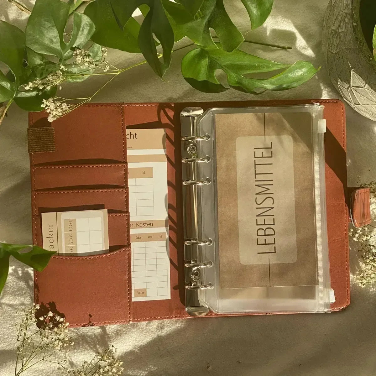 Eine Grundausstattung Budget Planung von Budget Binder by BeginnerBudget ist geöffnet und zeigt beschriftete Umschläge, Notizkarten und eine LEBENSMITTEL-Tasche, alles umgeben von Grünzeug und weißen Blumen im warmen Sonnenlicht.