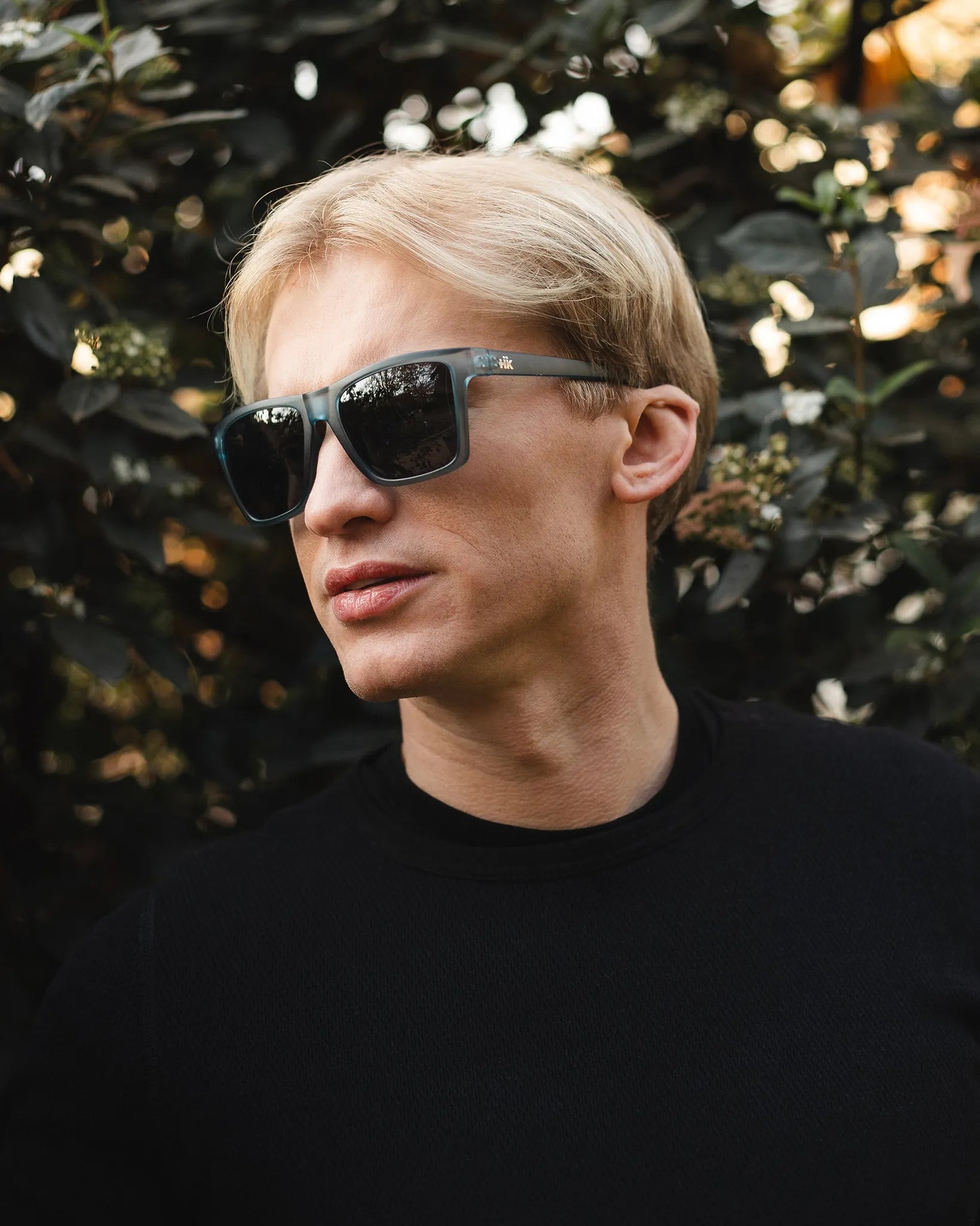 Eine Person mit kurzen blonden Haaren, die Sonnenbrille Breezy Blue Hanukeii by INDICOM BRANDS SL und ein schwarzes Hemd trägt, steht im Freien vor dunkelgrünem Laub.