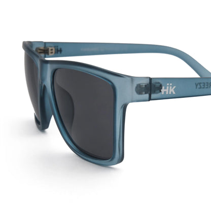 Nahaufnahme der Sonnenbrille Breezy Blue Hanukeii von INDICOM BRANDS SL, mit blauem Rahmen, dunklen Polycarbonat-Gläsern und weißem HK-Logo in der Nähe des Scharniers. Abgebildet auf einem sauberen weißen Hintergrund. UV400 Schutz sorgt für optimalen Augenschutz.