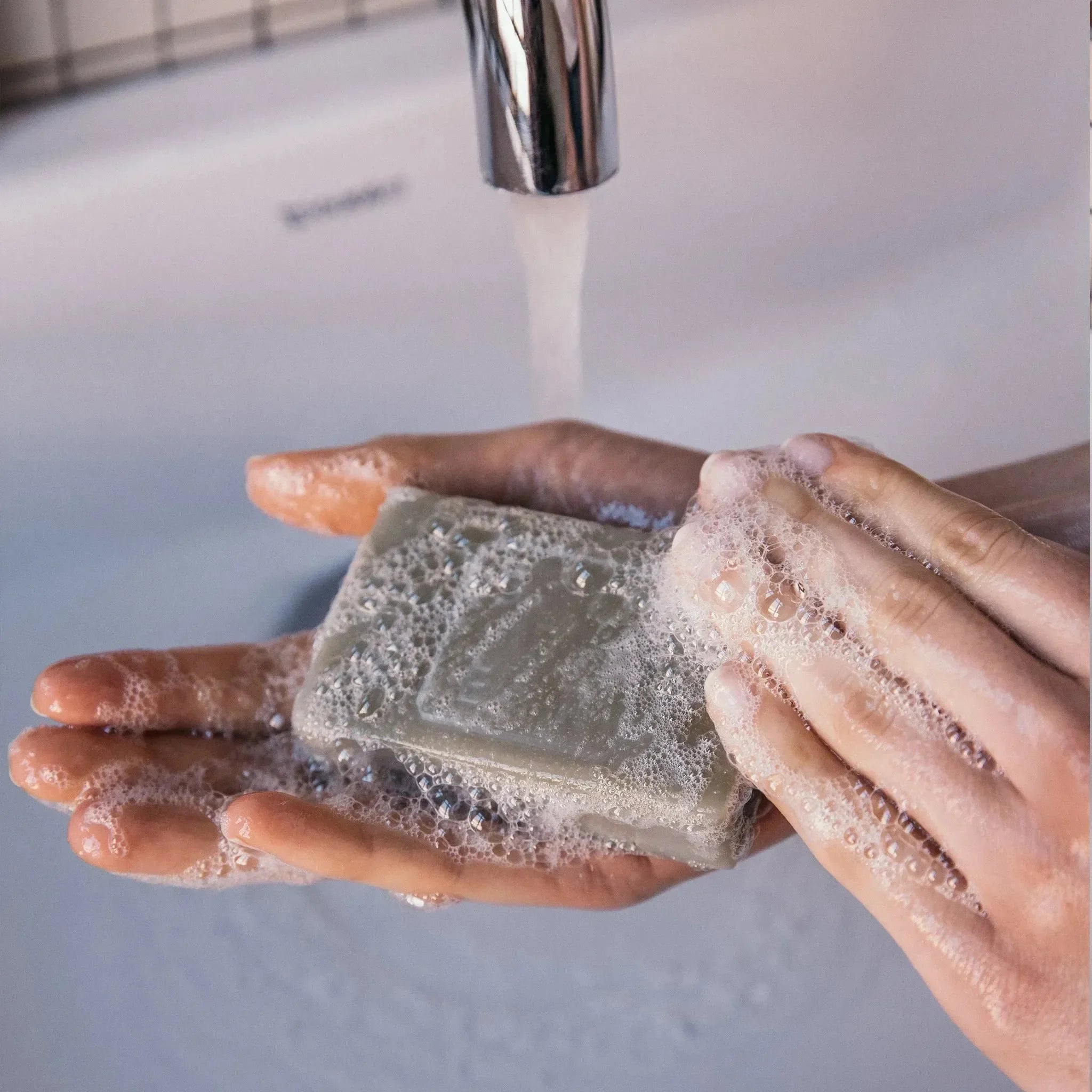 Nahaufnahme von Händen, die die nüchterne Hand & Body Moisturizer Soap Bar unter fließendem Wasser aufschäumen, wobei Schaum und Blasen über einem weißen Waschbecken entstehen.