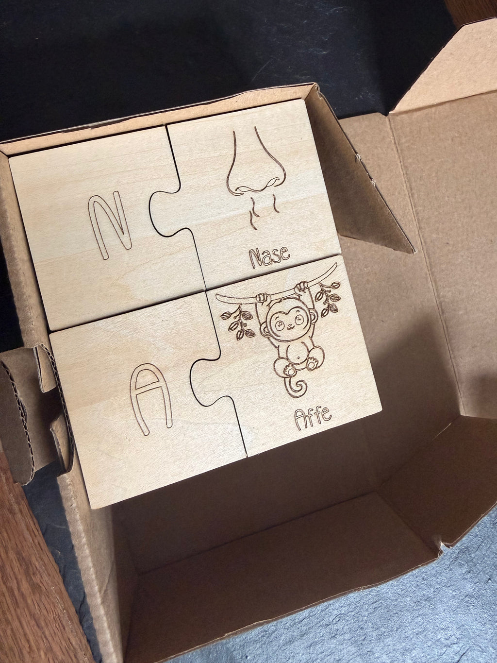 Wooden learning puzzle by flyliving with four pieces depicting a nose, a monkey, and letters 'N' and 'A' in a cardboard box.