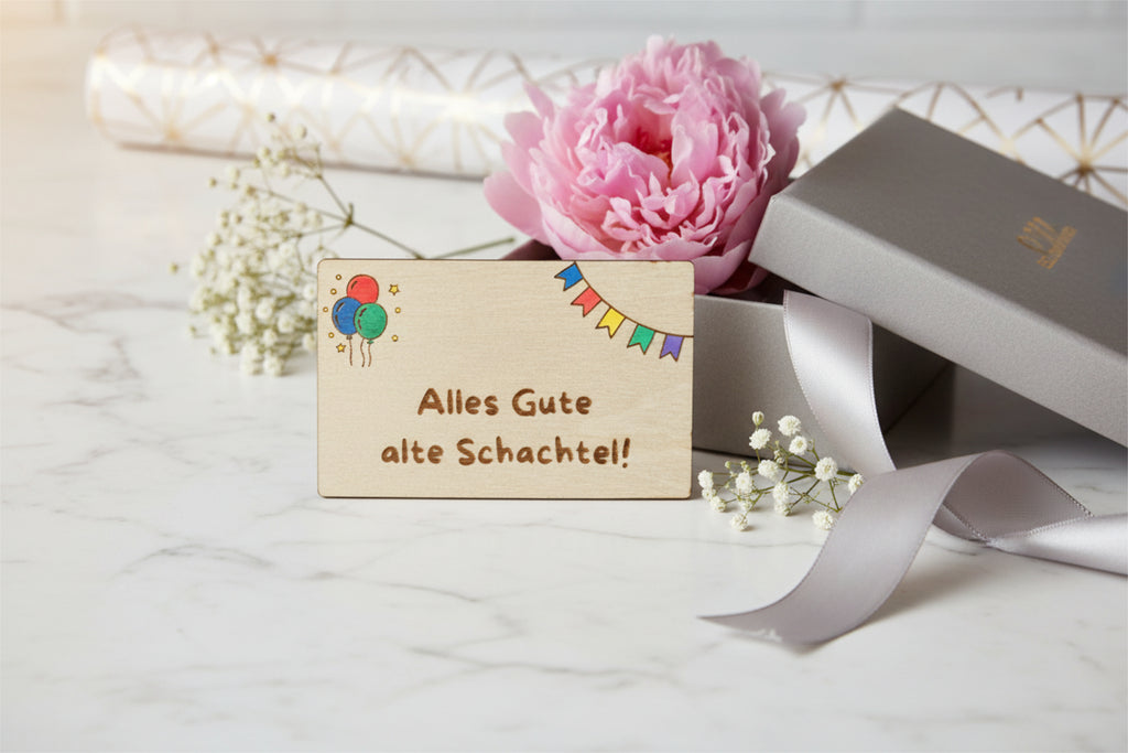 Wooden gift card by flyliving engraved with "Alles Gute alte Schachtel!", colorful balloons, and bunting, styled with a pink peony and silver gift box.