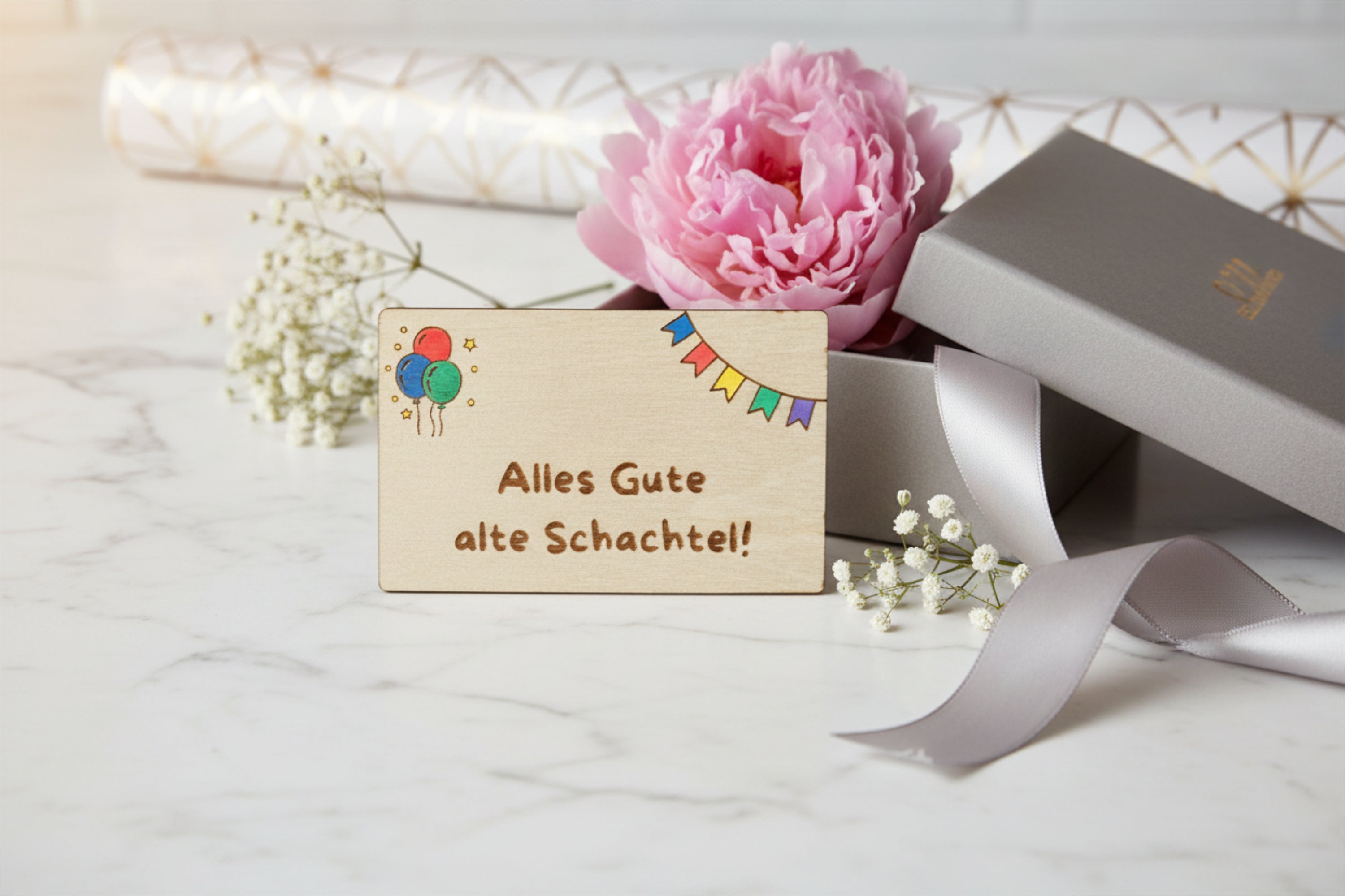 Wooden gift card by flyliving engraved with "Alles Gute alte Schachtel!", colorful balloons, and bunting, styled with a pink peony and silver gift box.