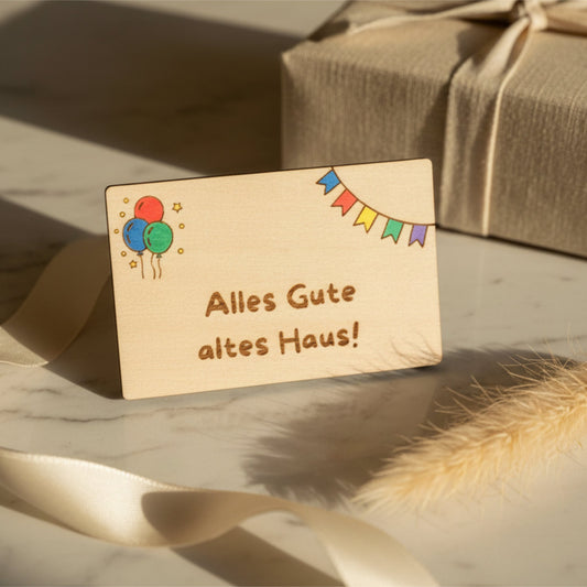 Light wooden gift card by flyliving engraved with "Alles Gute altes Haus!", colorful balloons, and bunting, resting on a marble surface.