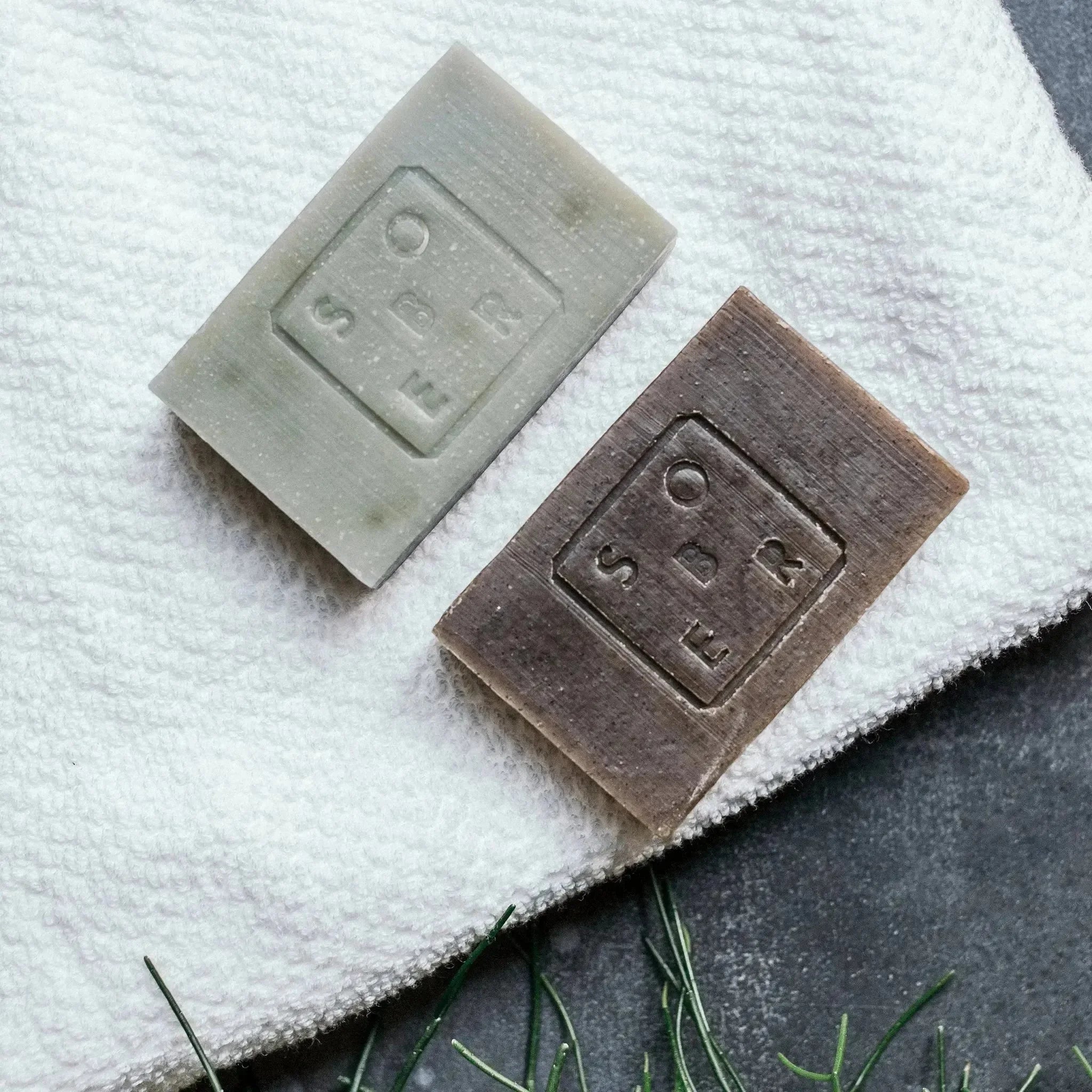 Zwei rechteckige, nüchterne Hand & Body Moisturizer Soap Bars, eine grüne und eine braune, liegen auf einem weißen, strukturierten Handtuch mit grünen Blättern in der Nähe, die auf einer dunklen Oberfläche ausgestellt sind.