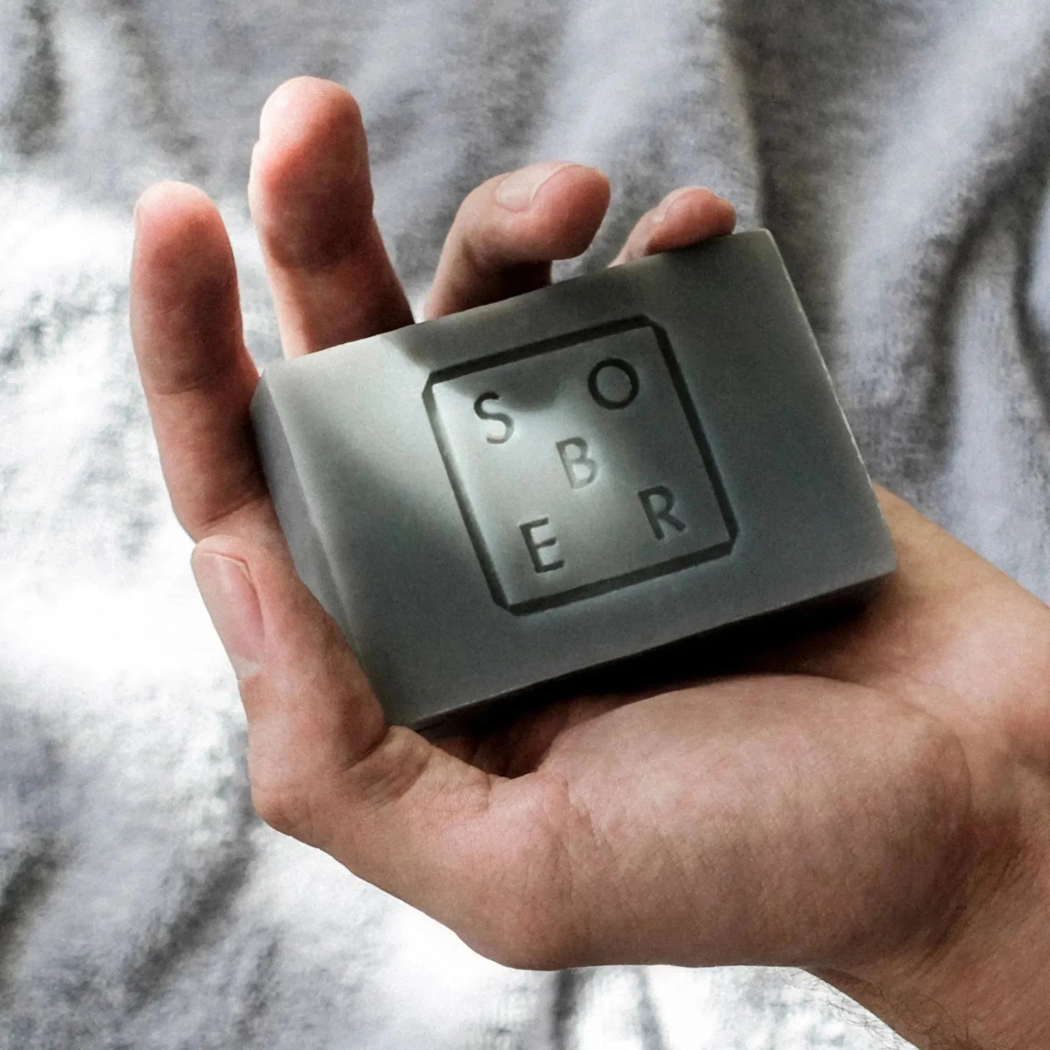 Eine Hand hält eine rechteckige Hand & Body Moisturizer Soap Bar von sober, auf deren Oberfläche das Wort "sober" eingraviert ist, vor einem verschwommenen, hellen Hintergrund.