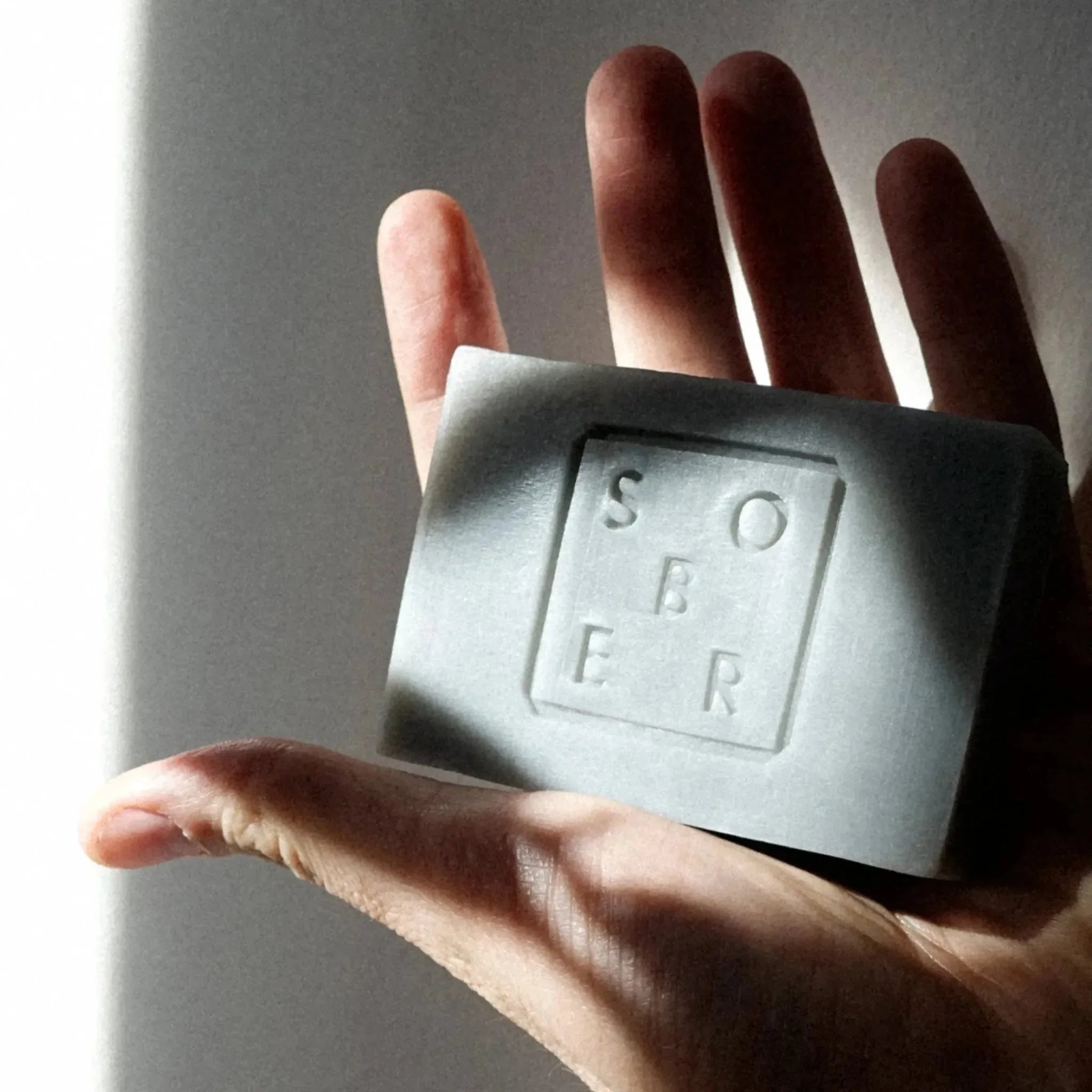 Eine Hand hält eine rechteckige Hand & Body Moisturizer Soap Bar von sober, auf der in einem Quadrat "sober" eingeprägt ist. Licht und Schatten beleuchten teilweise die Hand und die Seife.