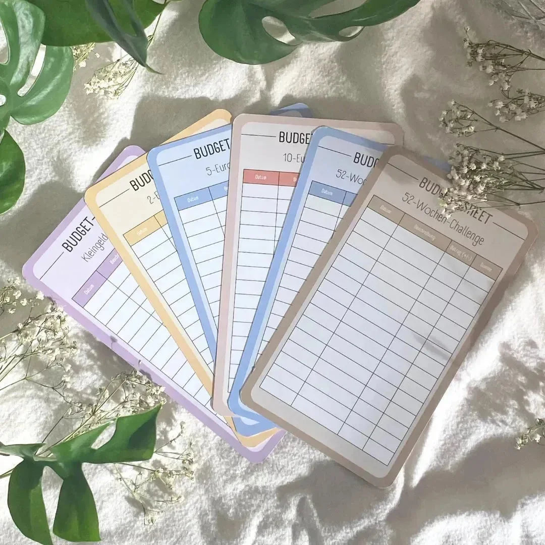 Sechs bunte Spar-Challenge-Set-Karten von BeginnerBudget sind auf einer weißen, strukturierten Oberfläche mit grünen Blättern und kleinen weißen Blumen aufgefächert. Jede Karte motiviert Sie mit einer anderen Spar-Challenge zum Sparen.