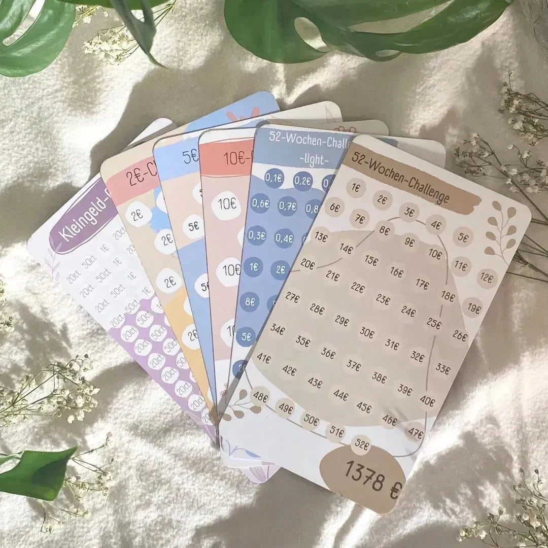 Fünf farbenfrohe Spar-Challenge-Set Karten von BeginnerBudget, für 52-wöchiges Sparen in Euro, sind auf einem strukturierten Stoff mit grünen Blättern und weißen Blumen abgebildet. Jede Karte zeigt wöchentliche Euro-Beträge zum Sparen und motiviert zum Geld sparen.