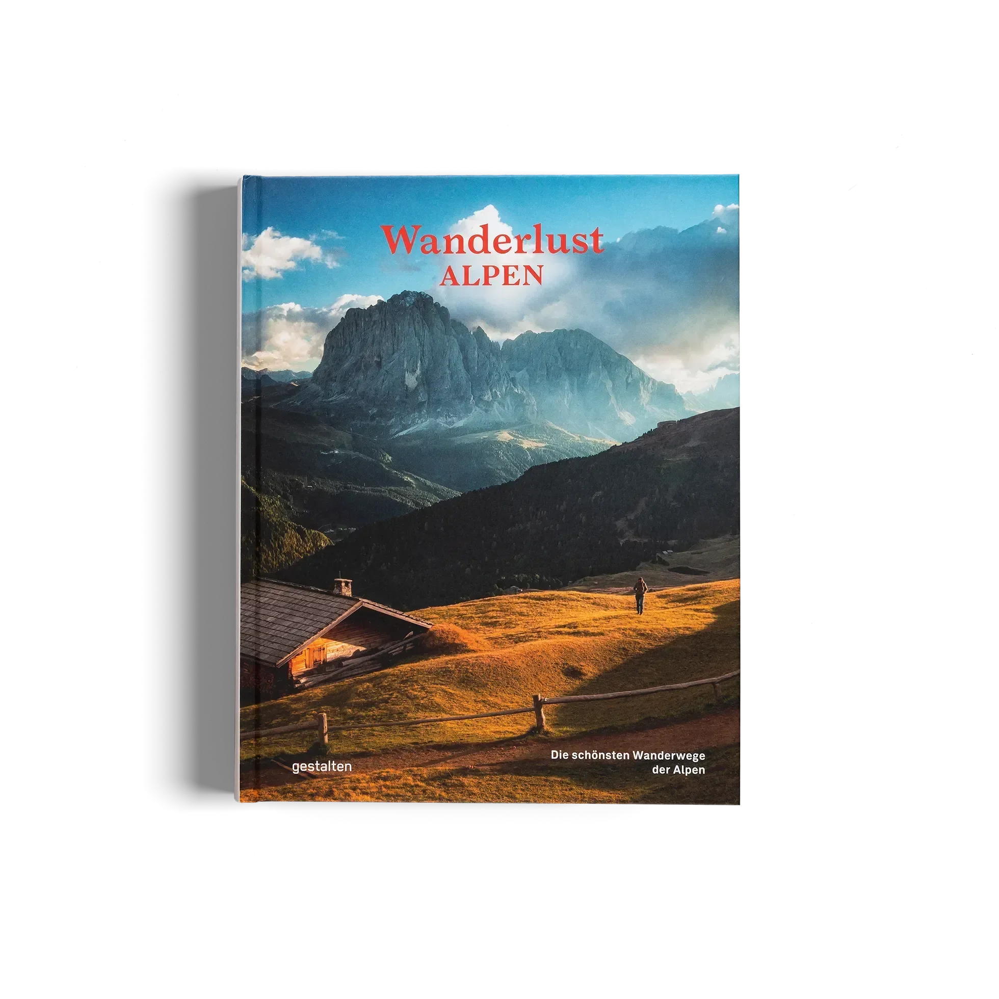 Wanderlust Alpen von gestalten EU Shop zeigt eine malerische Berglandschaft mit einer Hütte, grasbewachsenen Hügeln und einem Wanderer, der auf alpinen Pfaden unter einem blauen, wolkenverhangenen Himmel wandert.