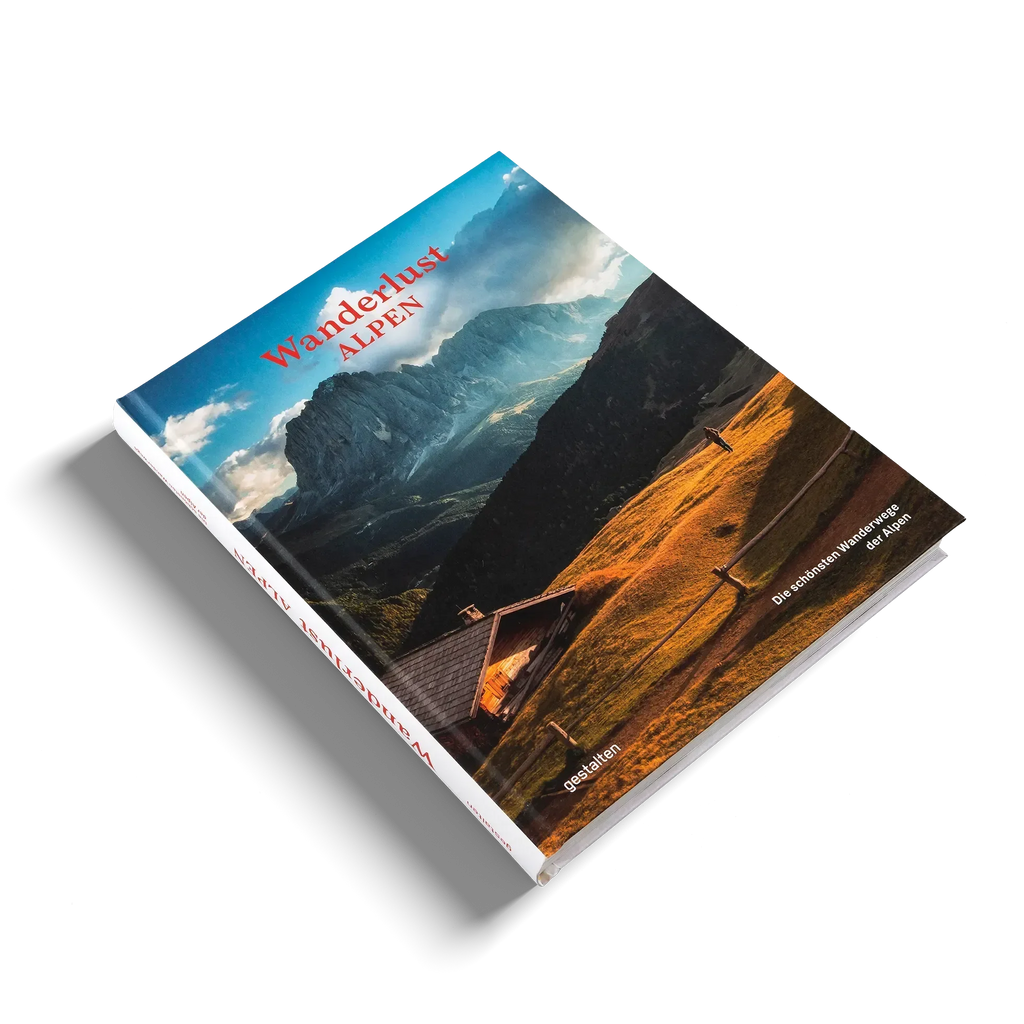 Das Buch "Wanderlust Alpen" von gestalten EU Shop liegt auf einer weißen Fläche und zeigt auf dem Cover eine Holzhütte inmitten goldener Hügel - ein Aufruf zu alpinen Abenteuern mit dramatischen Bergen und Wolken im Hintergrund.