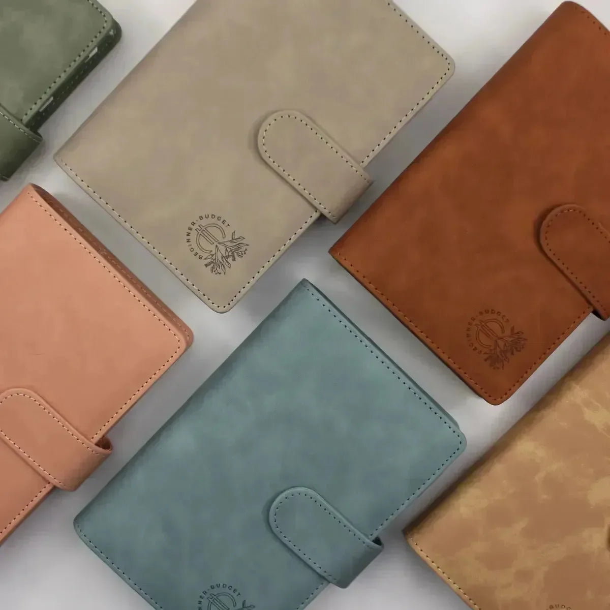 Sechs pastellfarbene Budget Binder (veganes) Wildleder - Personal (A6) von BeginnerBudget sind diagonal auf einem hellen Hintergrund angeordnet. Farben: grün, beige, braun, pfirsichfarben, blau und hellbraun; jede Farbe ist mit einem geprägten Logo versehen - ideal für eine stilvolle Finanzorganisation.