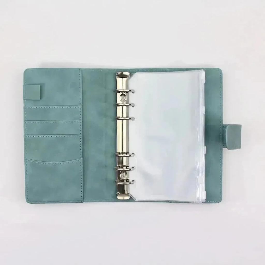 Der BeginnerBudget Budget Binder (veganes) Wildleder - Personal (A6) in teal verfügt über Metallringe mit Klarsichthüllen, Kartenfächer, eine Stiftschlaufe auf der linken Seite und eine Druckknopflasche auf der rechten Seite. Die lederähnliche Oberfläche ist auf einem schlichten weißen Hintergrund zu sehen.