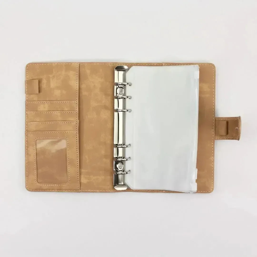 Der BeginnerBudget Budget Binder (veganes) Wildleder - Personal (A6) liegt aufgeklappt auf weißem Grund und zeigt links Metallringe mit Klarsichthüllen für die Finanzübersicht, Kartenschlitze und Ausweisfenster und rechts eine Druckknopf- oder Magnetverschlusslasche.