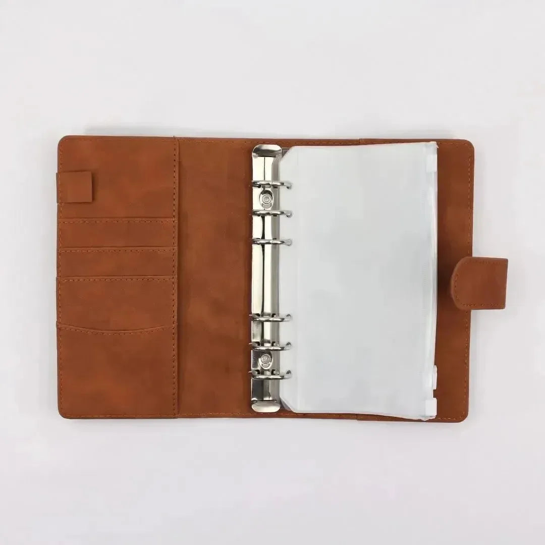 Der BeginnerBudget Budget Binder (veganes) Wildleder - Personal (A6) hat ein offenes Design aus braunem veganem Wildleder mit Metallringen, Kartenfächern auf der linken Seite und durchsichtigen Plastikhüllen auf der rechten Seite, dargestellt auf einem schlichten weißen Hintergrund.