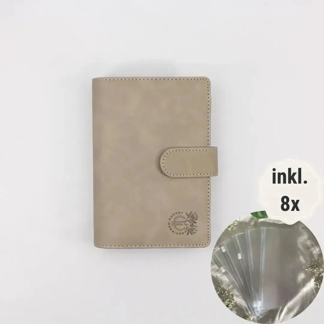 Ein grauer Budget Binder (A6) aus veganem Wildleder von BeginnerBudget mit Druckknopfverschluss liegt geschlossen auf einem weißen Hintergrund. Ein Einschub zeigt 8 mitgelieferte Klarsichthüllen für Sparen oder bessere Finanzübersicht, beschriftet mit "inkl.". 8x".
