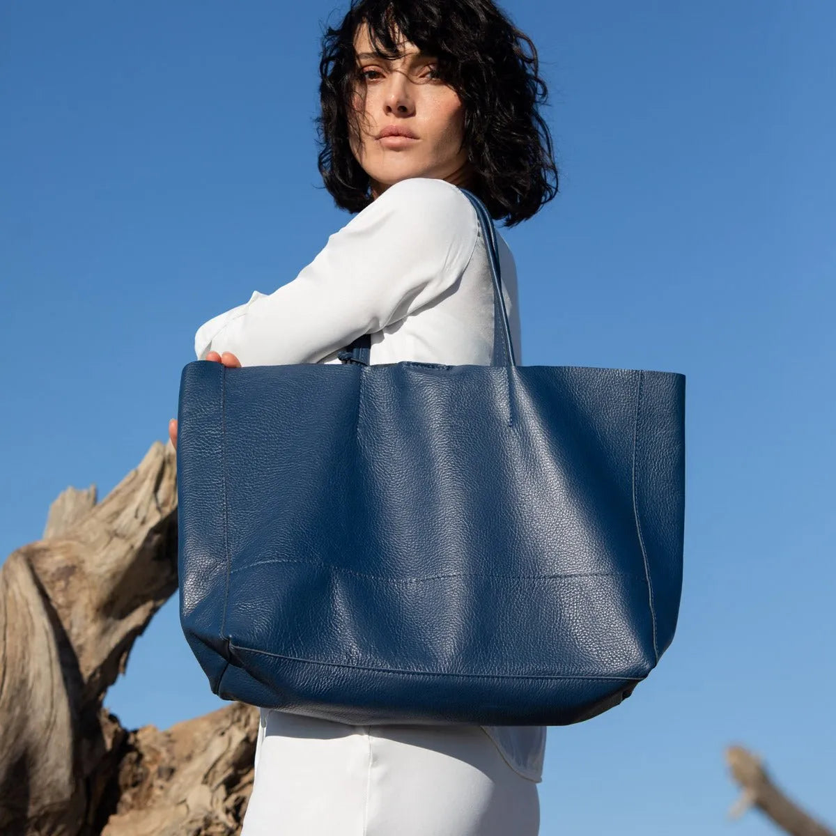 Eine Frau mit kurzen dunklen Haaren in weißem Outfit steht im Freien vor einem klaren blauen Himmel und trägt die marineblaue Umhängetasche "Susi" aus Leder von flyliving über ihre Schulter.