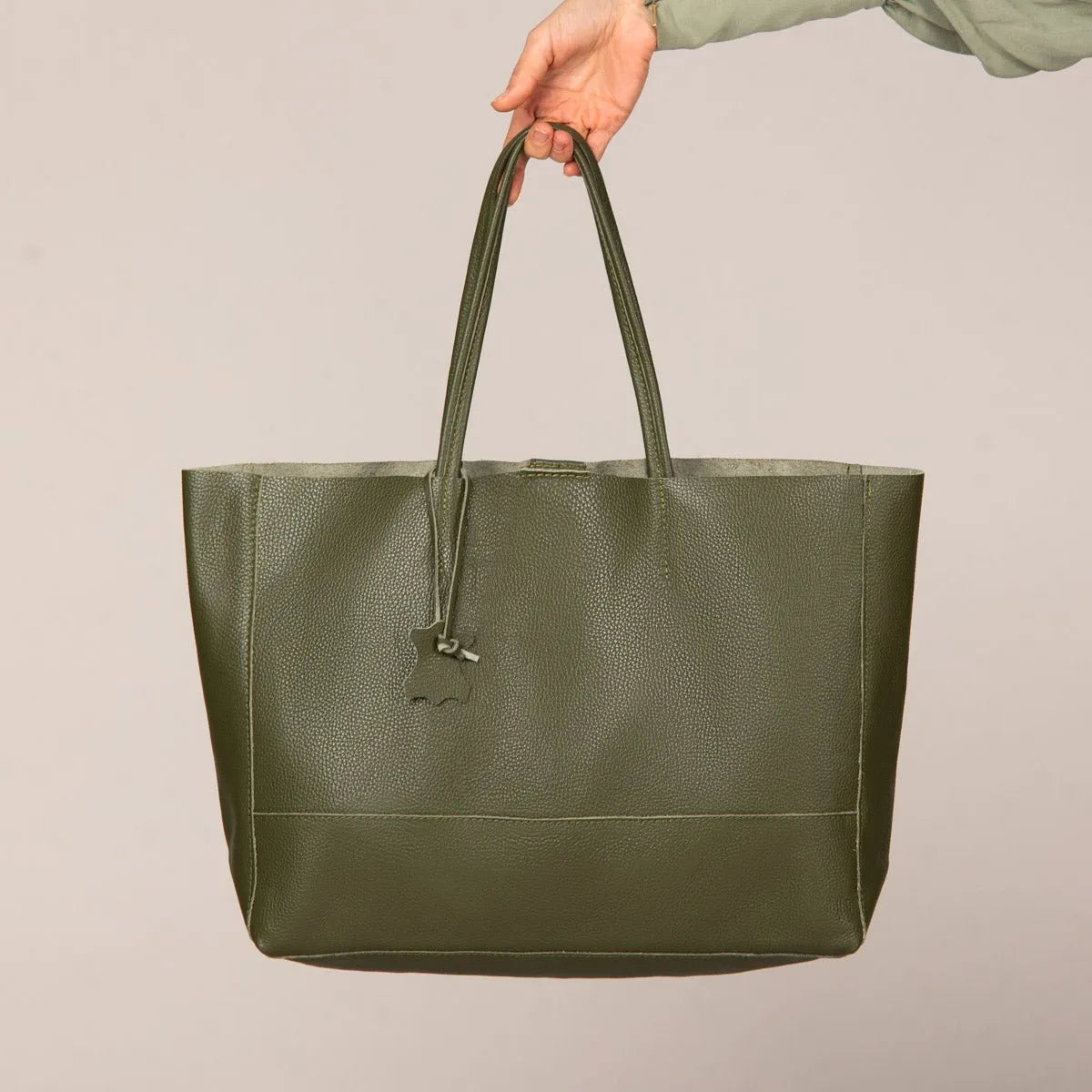 Eine Hand hält die grüne flyliving Umhängetasche "Susi" aus Leder, mit Doppelgriffen und einem kleinen Anhänger, vor einem schlichten beigen Hintergrund.